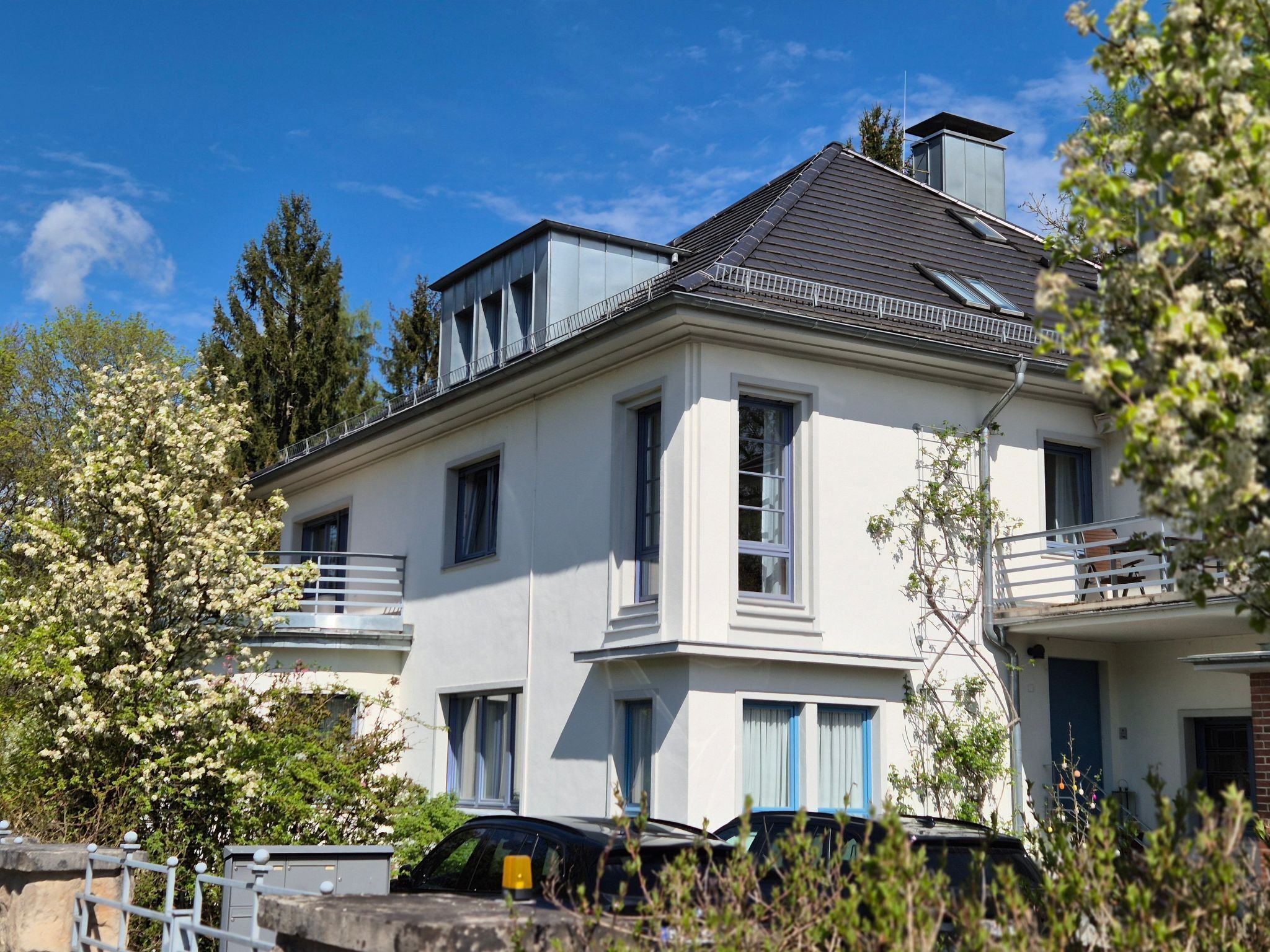A two-story house with a balcony, a car parked in front, and a tree with white flowers.