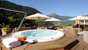 Ein Whirlpool mit Holzterrasse und Sitzgelegenheiten, umgeben von Sonnenschirmen, mit Blick auf eine gebirgige Landschaft.