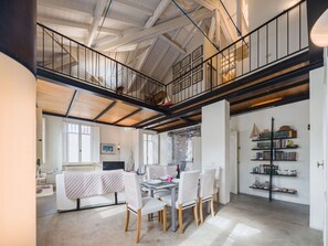 Un loft amplio con techo alto, vigas a la vista, un comedor con mesa de vidrio y una biblioteca con estanterías.