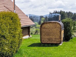 Eine Holzhütte mit Schornstein, aus dem Rauch aufsteigt, inmitten einer üppigen, grünen Landschaft und einem Haus mit rotem Dach.