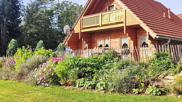 Una casa de madera con balcón, rodeada de un jardín con diversas plantas y flores.