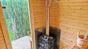 Interior de una cabaña de madera con chimenea central, una puerta que da al exterior y un banco de madera.