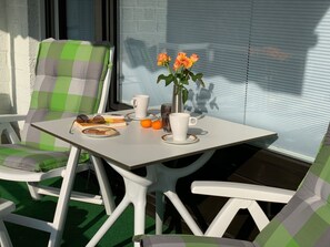 Ein Tisch mit einer weißen Tasse, einer weißen Kaffeetasse, einem Buch und einem Teller mit Keksen. Zwei orangefarbene Früchte stehen auf dem Tisch. Eine Vase mit Blumen steht ebenfalls auf dem Tisch.