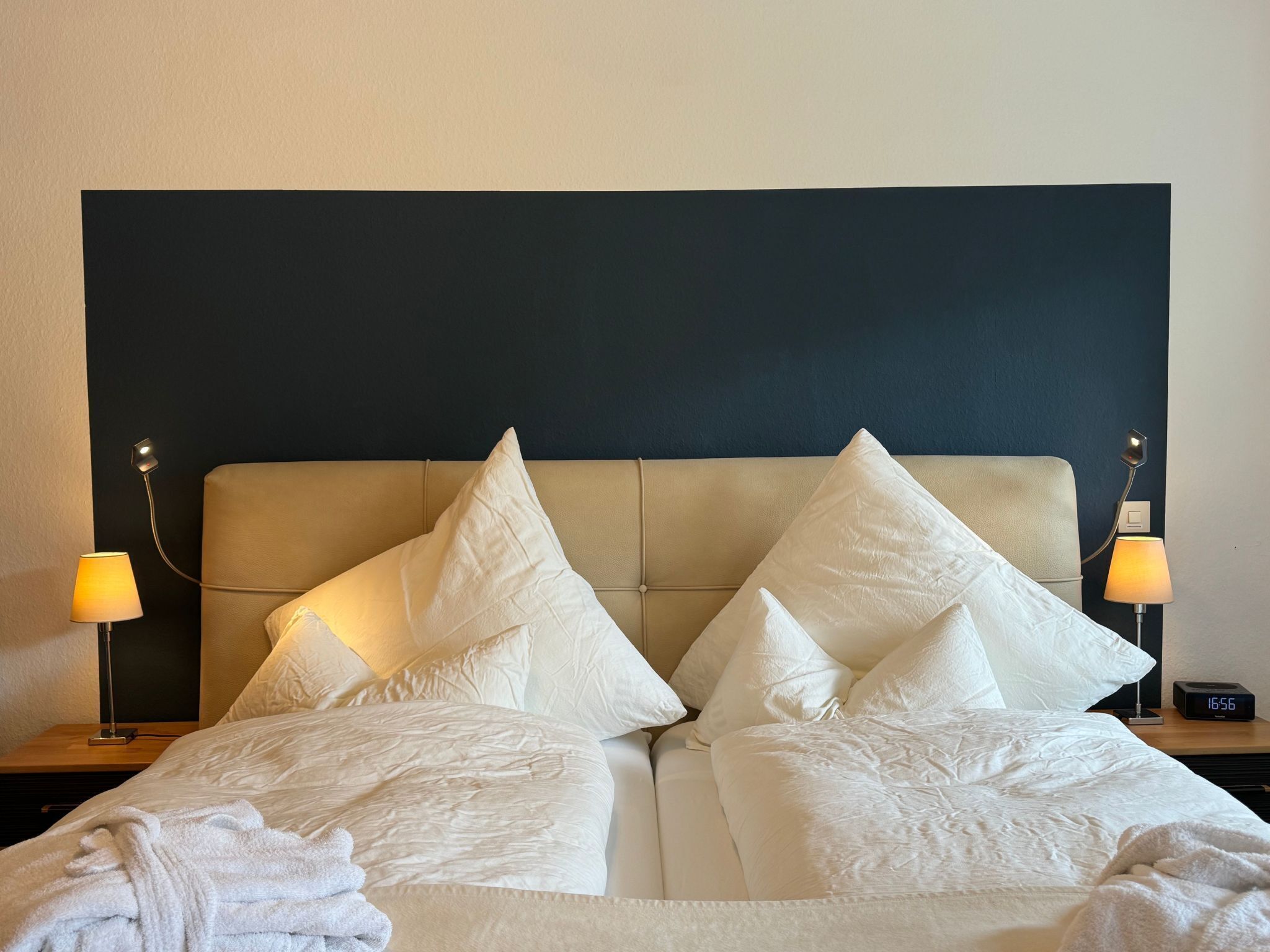 A neatly made bed with white linens and pillows, two bedside lamps, and a digital clock on a bedside table.