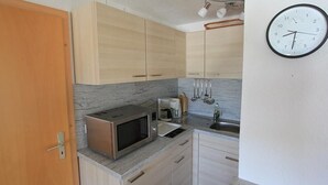Een compacte keuken met houten kastjes, een magnetron, een spoelbak en een klok aan de muur.