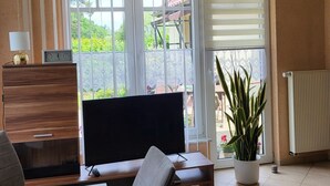 Una sala de estar con televisor, una mesa de comedor, sillas, una planta en maceta y vistas al exterior a través de amplios ventanales.