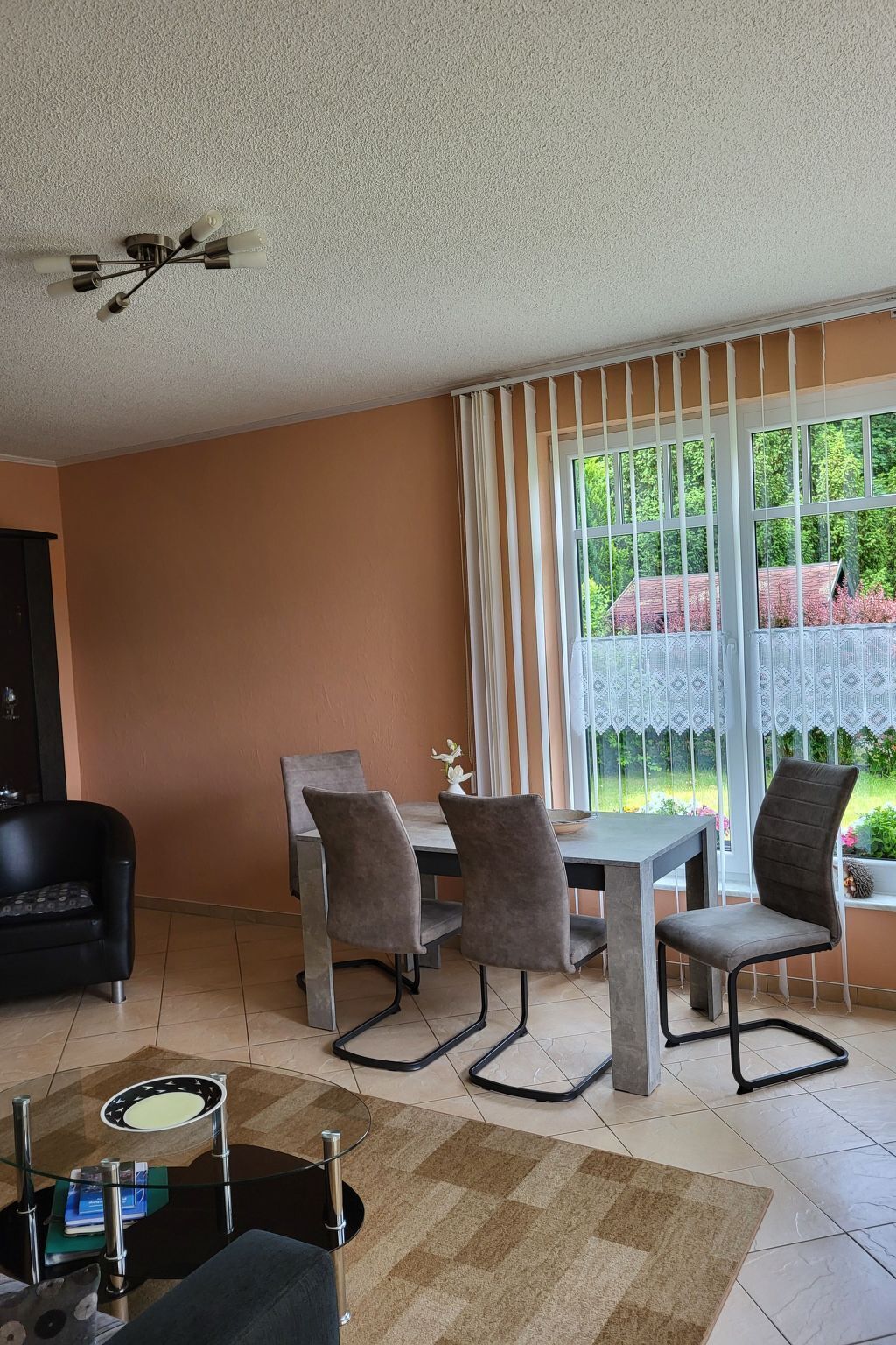 A dining area with a table and chairs, a sofa, a glass coffee table, and a view of greenery through large windows.