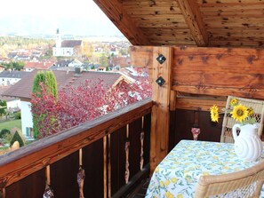 Ein Balkon mit Holzgeländer, ein Korbstuhl, ein Tisch mit blumigem Tischläufer und eine Vase mit Sonnenblumen.