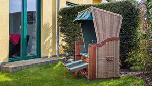 Uma área de estar ao ar livre com mobiliário de vime, um toldo verde, um banco de madeira e uma pequena mesa, localizada ao lado de um edifício com fachada amarela e porta verde.