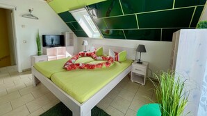 Un dormitorio con techo inclinado, cama con ropa de cama verde, televisor sobre un mueble y ventana con vistas.