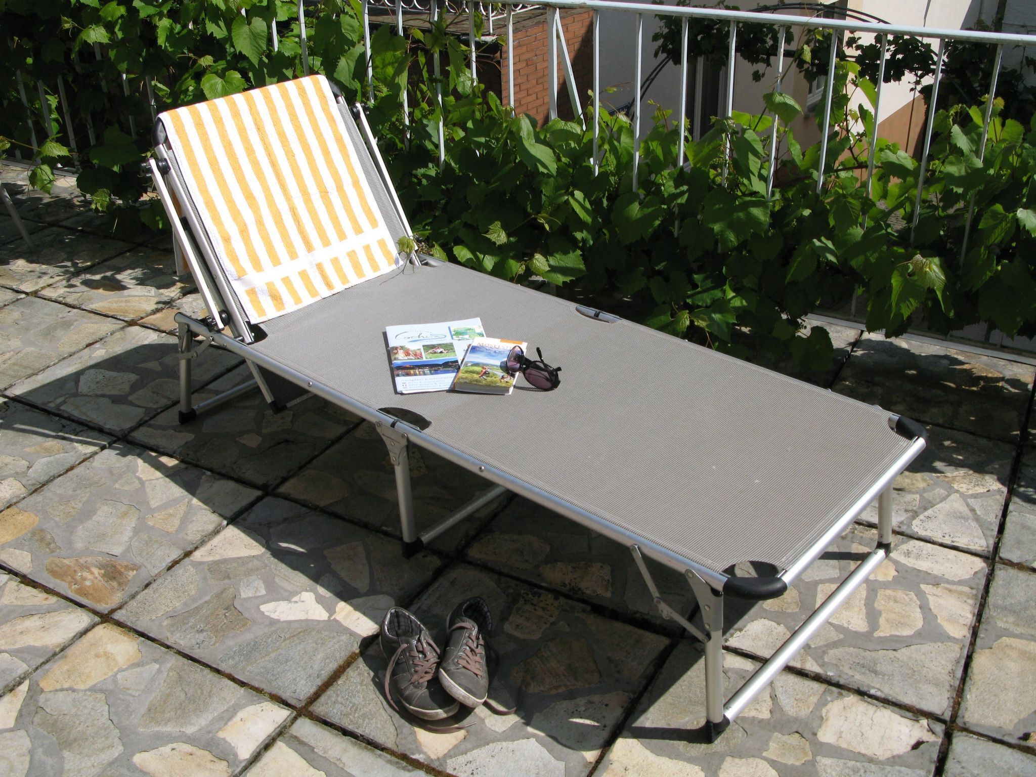 Une chaise longue en métal avec une serviette à rayures, une paire de chaussures et un magazine, le tout posé sur une terrasse en pierre.