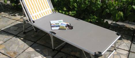 Une chaise longue en métal avec une serviette à rayures, une paire de chaussures et un magazine, le tout posé sur une terrasse en pierre.