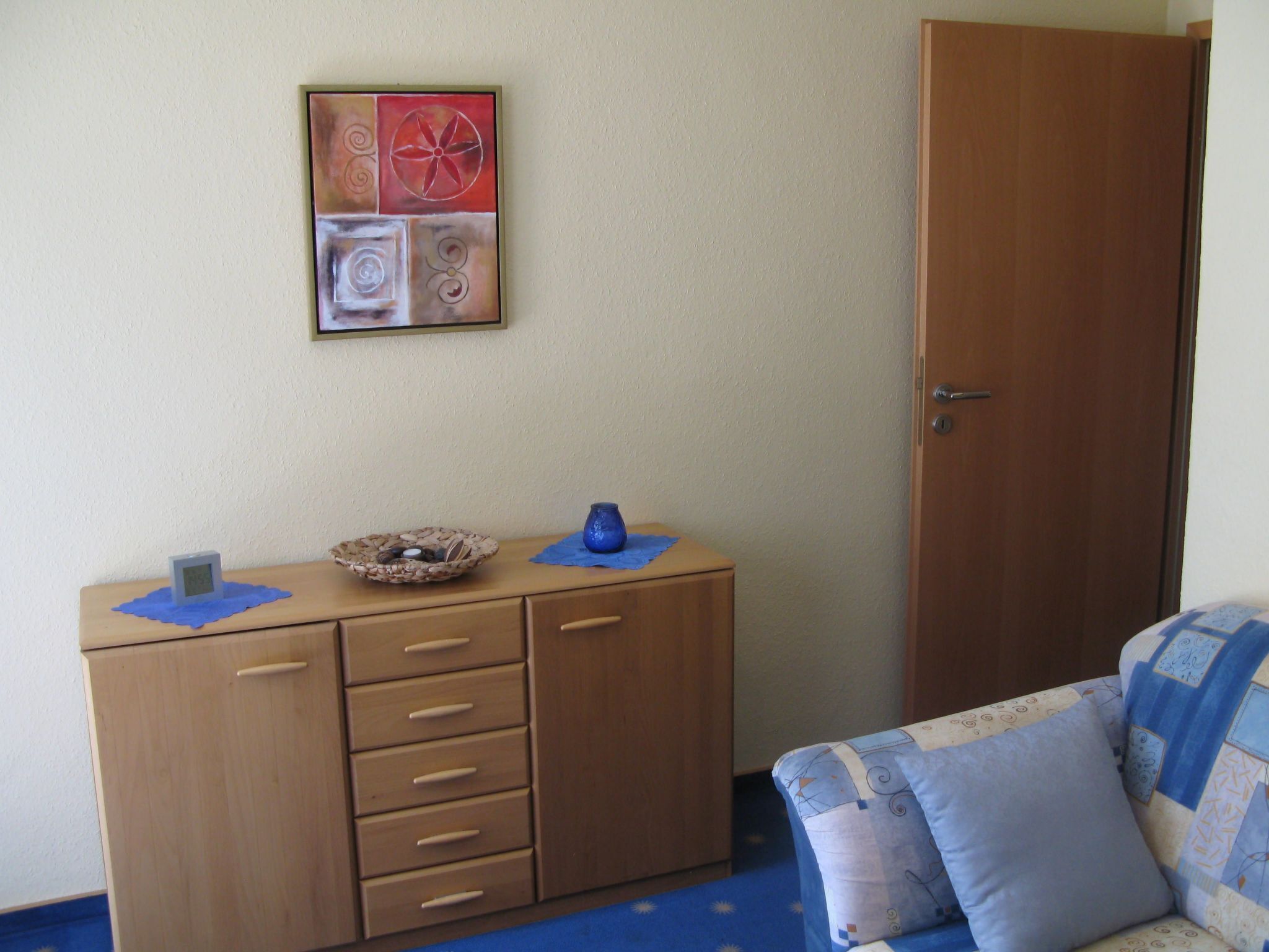A room with a wooden dresser, a blue cushion, and a wall-mounted abstract painting.
