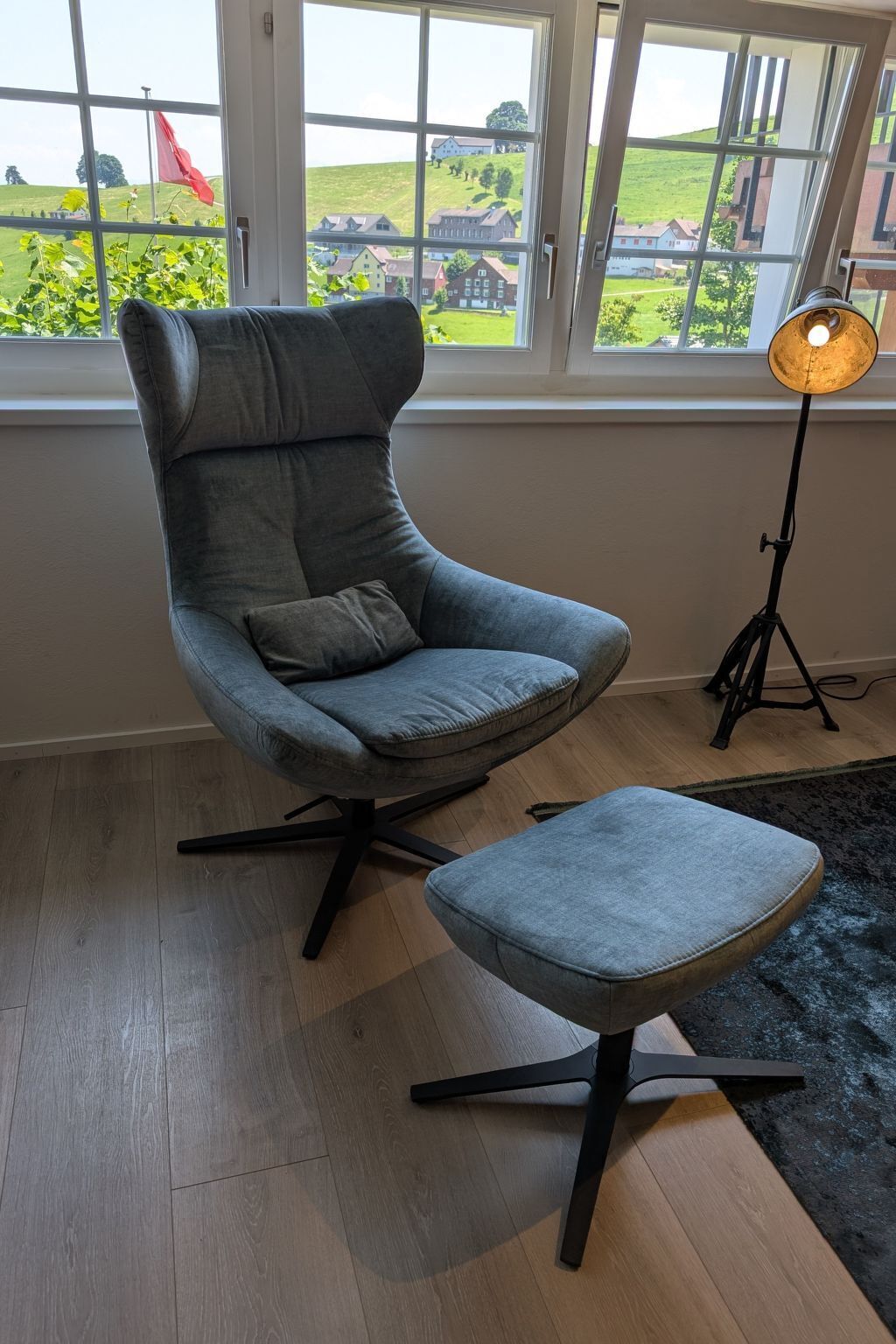 Un sillón moderno gris con su otomana a juego, ubicado frente a un ventanal con vistas a un paisaje verde y edificios.