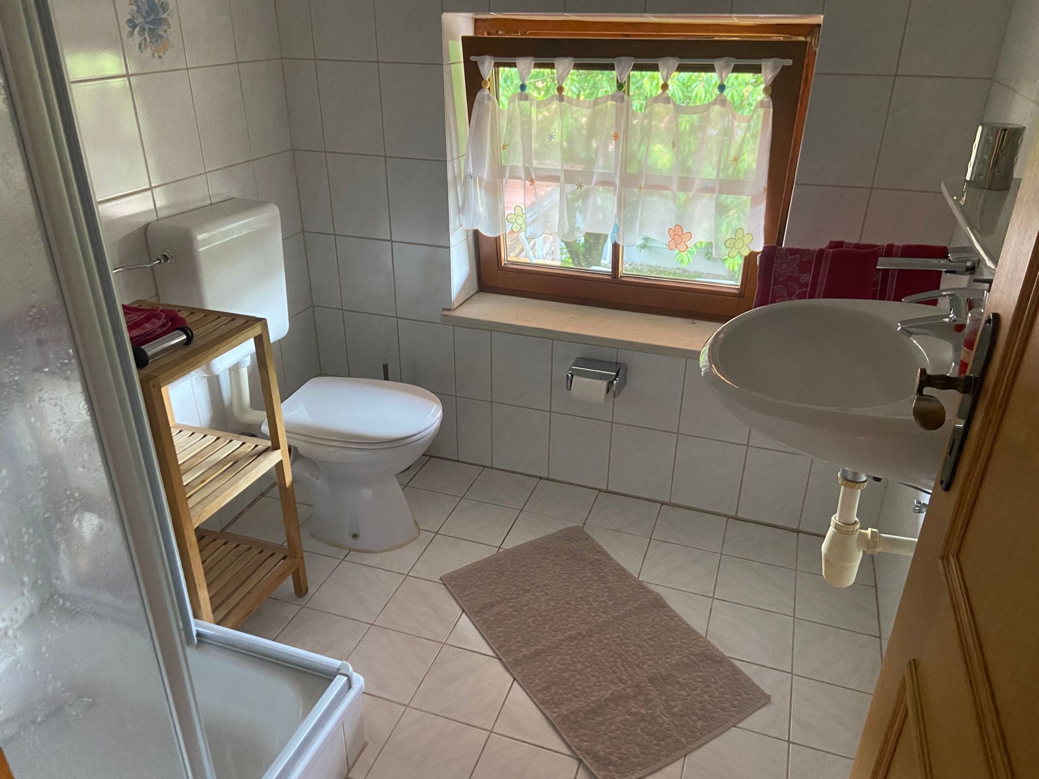 A small bathroom with a toilet, sink, window, and a wooden shelf.