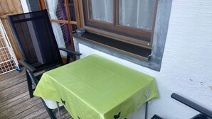 A small outdoor table with a green tablecloth, a black chair, and a window with a wooden frame and white curtains.