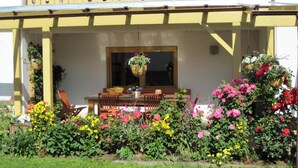 Un edificio amarillo con un porche cubierto, una mesa de comedor con sillas y una cesta de flores colgante.
