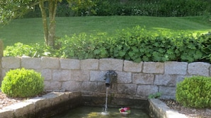 A stone fountain with water spouting from a central pipe, surrounded by a low stone wall and bordered by neatly trimmed bushes. - Tauchert by Interhome (Baiersbronn)