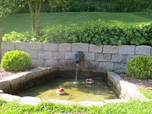 Ein Steinfbrunnen, aus dessen mittlerem Rohr Wasser sprudelt, umgeben von einer niedrigen Steinmauer und gesäumt von ordentlich geschnittenen Büschen.