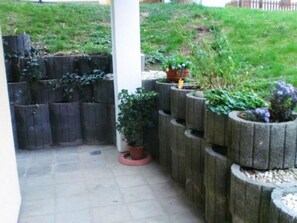 Un espace jardin aménagé avec plusieurs jardinières disposées en terrasses, une colonne blanche et une pente herbeuse en arrière-plan.