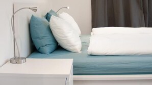 Een slaapkamer met een bed, kussens, een nachtkastje, een lamp, een raam met gordijnen en een schilderij aan de muur.