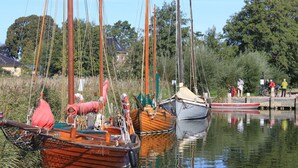 Un cours d’eau paisible où plusieurs bateaux sont amarrés, entouré de verdure et où quelques personnes se trouvent près du rivage.