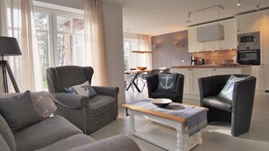 Un salon moderne avec des canapés gris, une table basse et un coin repas comprenant une table et des chaises. On aperçoit, au fond, une cuisine avec des meubles blancs et des appareils électroménagers.