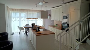 Une cuisine moderne avec des meubles blancs, un îlot central, un coin repas avec des chaises et un escalier avec des rampes blanches.