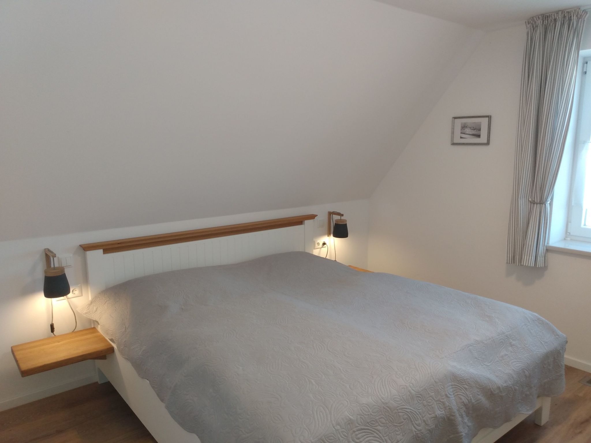 Een slaapkamer met een bed, een houten hoofdbord, bedlampen, een klein plankje en een raam met gordijnen.