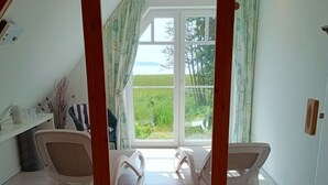 Une pièce avec un plafond haut, un banc en bois, deux chaises blanches, une petite table et une grande fenêtre avec des rideaux.