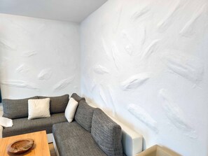 En hörna i ett rum med en grå soffa, ett bord i trä och en vit vägg med ett mönstrat ytskikt.
