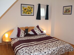Una camera da letto con soffitto spiovente, un letto con copriletto decorato, due quadri appesi alle pareti e una finestra con tende.
