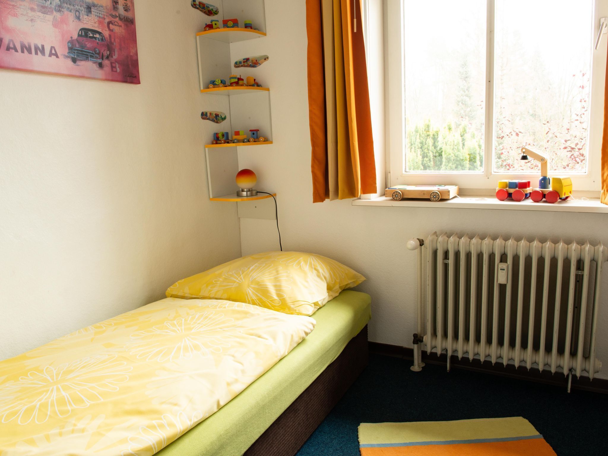 Une petite chambre avec un lit simple, un radiateur, une fenêtre avec des rideaux et une étagère remplie de jouets.
