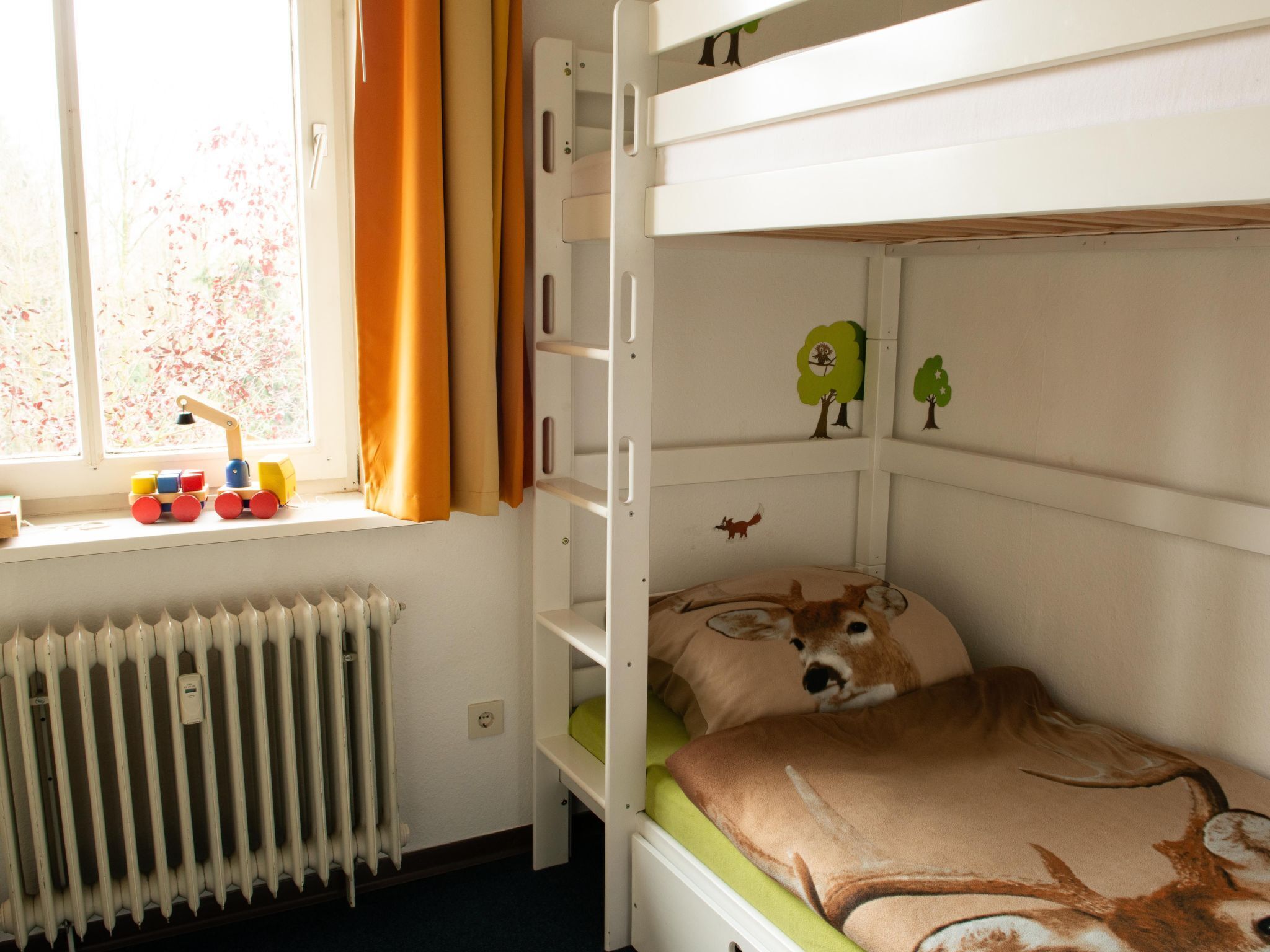 A bunk bed with a green mattress and a pillow with a deer design. A toy train is on the windowsill, and there are wall decorations of trees and animals.