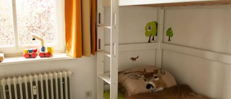 A bunk bed with a green mattress and a pillow with a deer design. A toy train is on the windowsill, and there are wall decorations of trees and animals.