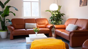 Ein Wohnzimmer mit zwei braunen Ledersofas, einem weißen Couchtisch und einer gelben Decke. Ein großes Fenster lässt viel Tageslicht herein, daneben ein Blumentopf und eine Pendelleuchte.