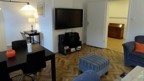 Uma sala de estar com televisão de tela plana, uma mesa de centro, um sofá e um espaço de jantar com mesa e cadeiras.