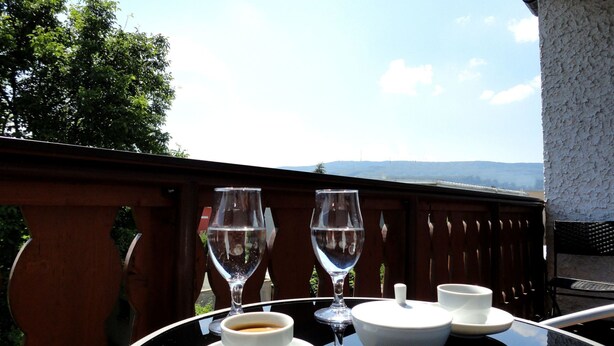 A table with a cup of coffee, a glass of water, and a wine glass on a balcony with a view of the mountains.