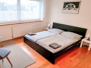 Un dormitorio con cama, una silla, una mesita de noche, una lámpara, una ventana y un cuadro en la pared.