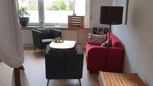 Un accogliente soggiorno con un divano rosso, una poltrona nera, un tavolino e un tavolino di vimini. Si vede una finestra con tende decorate e una lampada a sospensione.