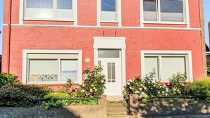 Un edificio a due piani con una facciata rossa, finestre con cornice bianca e un portone centrale, affiancato da un piccolo giardino con fiori gialli.