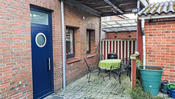 Un edificio in mattoni con una porta blu, una piccola zona esterna con un tavolo e sedie, e un cestino verde.