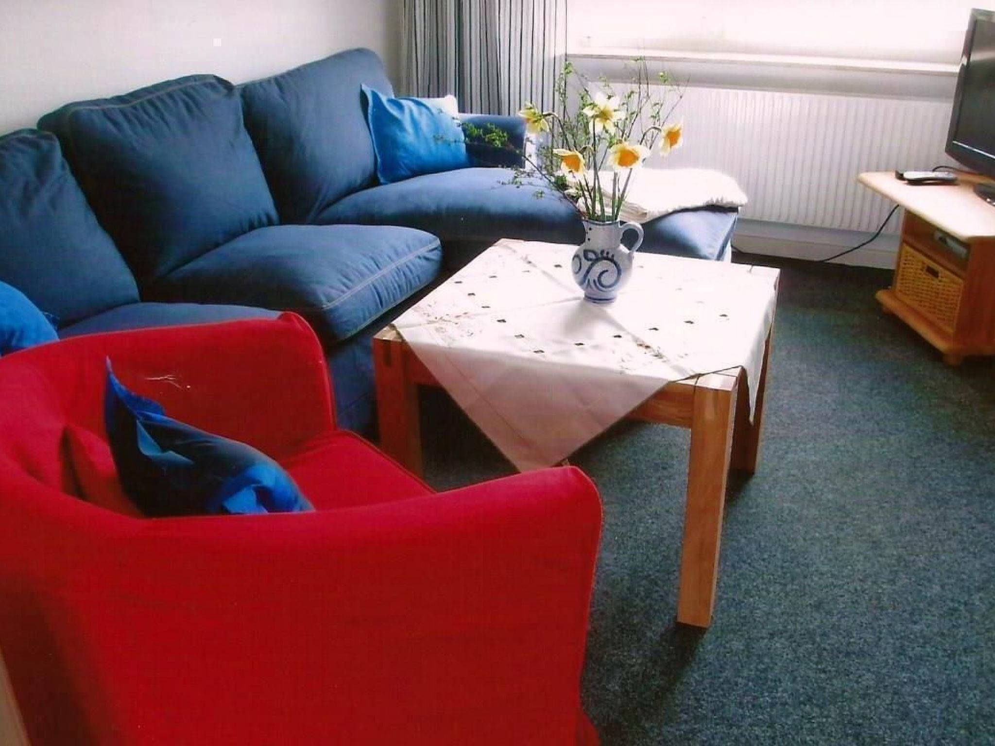 Ett vardagsrum med en blå soffa, en röd fåtölj och ett litet bord med en vas med blommor.