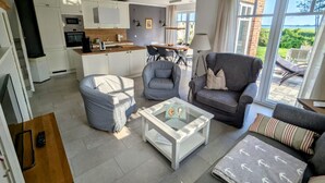 A modern living room with a sofa, armchair, and coffee table, a kitchen with white cabinets, and a view of the outdoors through large windows. - Admiral's Corner Luv by Interhome (Born)