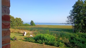 A view of a grassy field with a body of water in the background, a brick wall on the left, and some trees and shrubs in the foreground. - Admiral's Corner Luv by Interhome (Born)