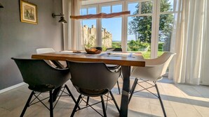 A dining area with a wooden table, two black chairs, and one white chair. There is a large window with curtains, a picture on the wall, and a hanging lamp. - Admiral's Corner Luv by Interhome (Born)