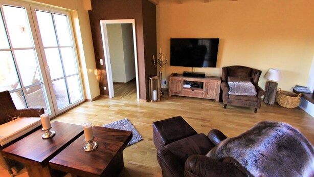 Een woonkamer met een houten plafond, een flatscreen televisie, een donkerbruine bank, een houten salontafel en een groot raam met jaloezieën.