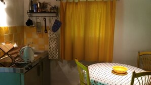 A small kitchen with a stove, kettle, and a table with a yellow bowl. There are chairs around the table and a yellow curtain. - weg von der Zuvielisation by Interhome (Menzonio)