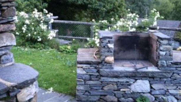Un caminetto esterno in pietra circondato da un muro di pietra, situato in un giardino con le montagne sullo sfondo.