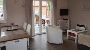 Une pièce comprenant une table à manger, des chaises, un petit meuble blanc, un téléviseur posé sur un meuble, et une porte-fenêtre coulissante donnant sur un espace extérieur.